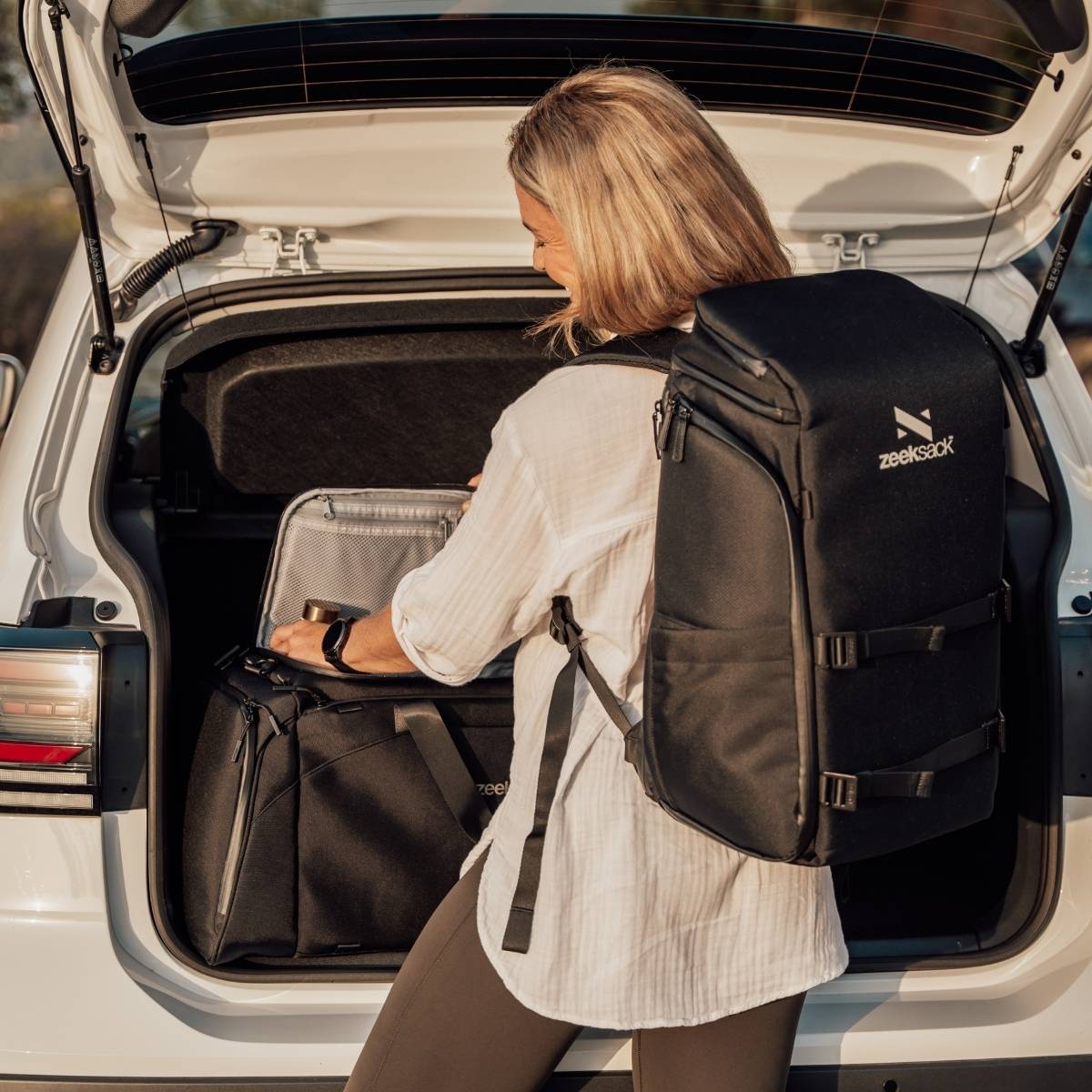 Woman wearing a black backpack next to an open car trunk.